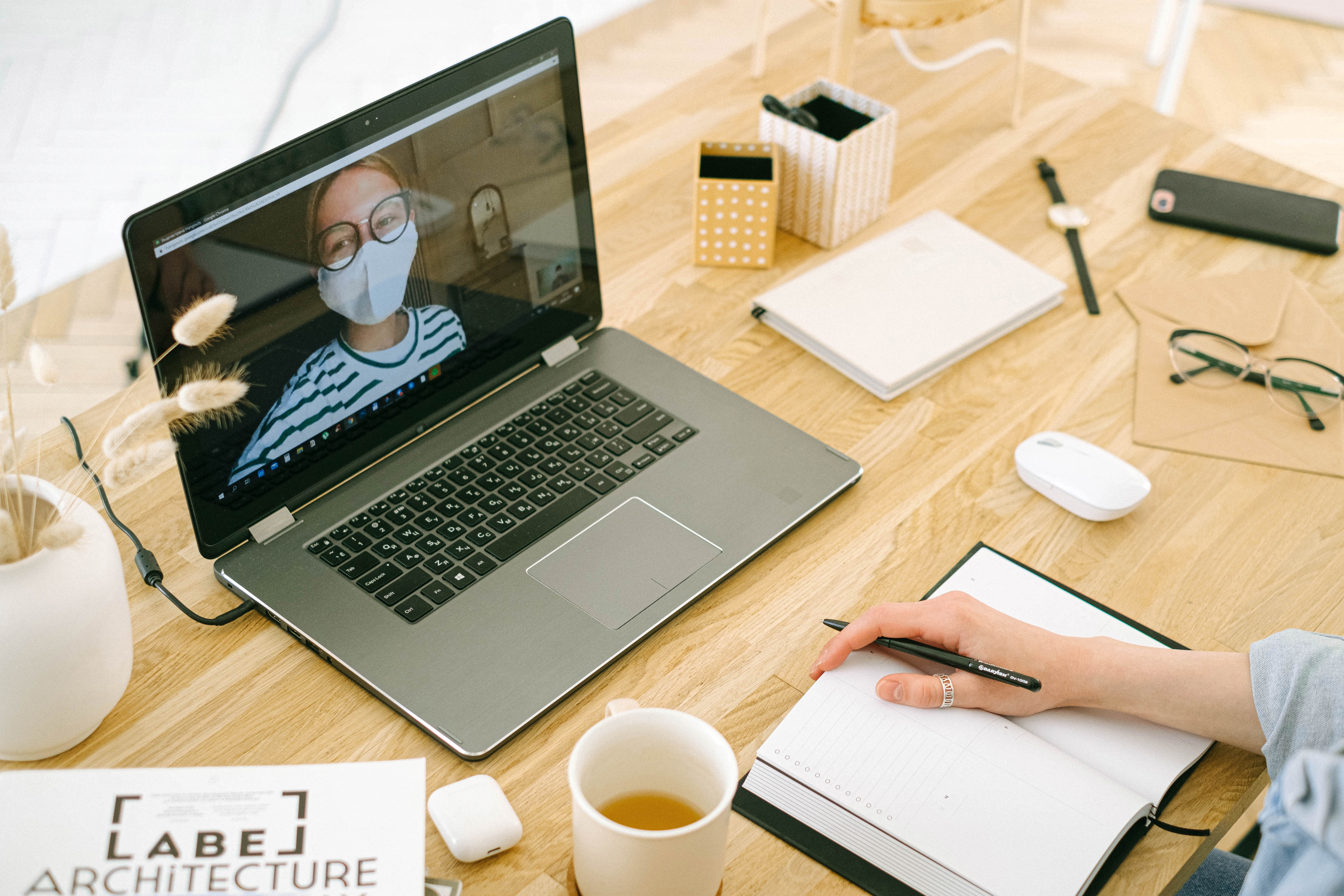  Online consultation with a legal professional, featuring a person wearing a mask on a laptop screen, surrounded by a well-organized desk with notepads, a cup of coffee, and office supplies, highlighting remote legal services during COVID-19.