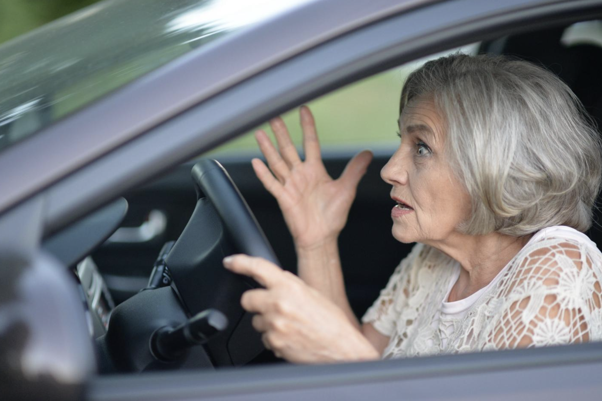 An older woman appears startled or confused while sitting behind the wheel of a car, her expression and gesturing hand indicating she may be reacting to a driving situation or potential traffic incident.