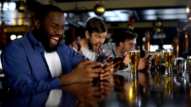 Group of friends at a bar, excitedly using smartphones with pints of beer on the bar counter, possibly engaging in legal sports betting activities available in Arizona.