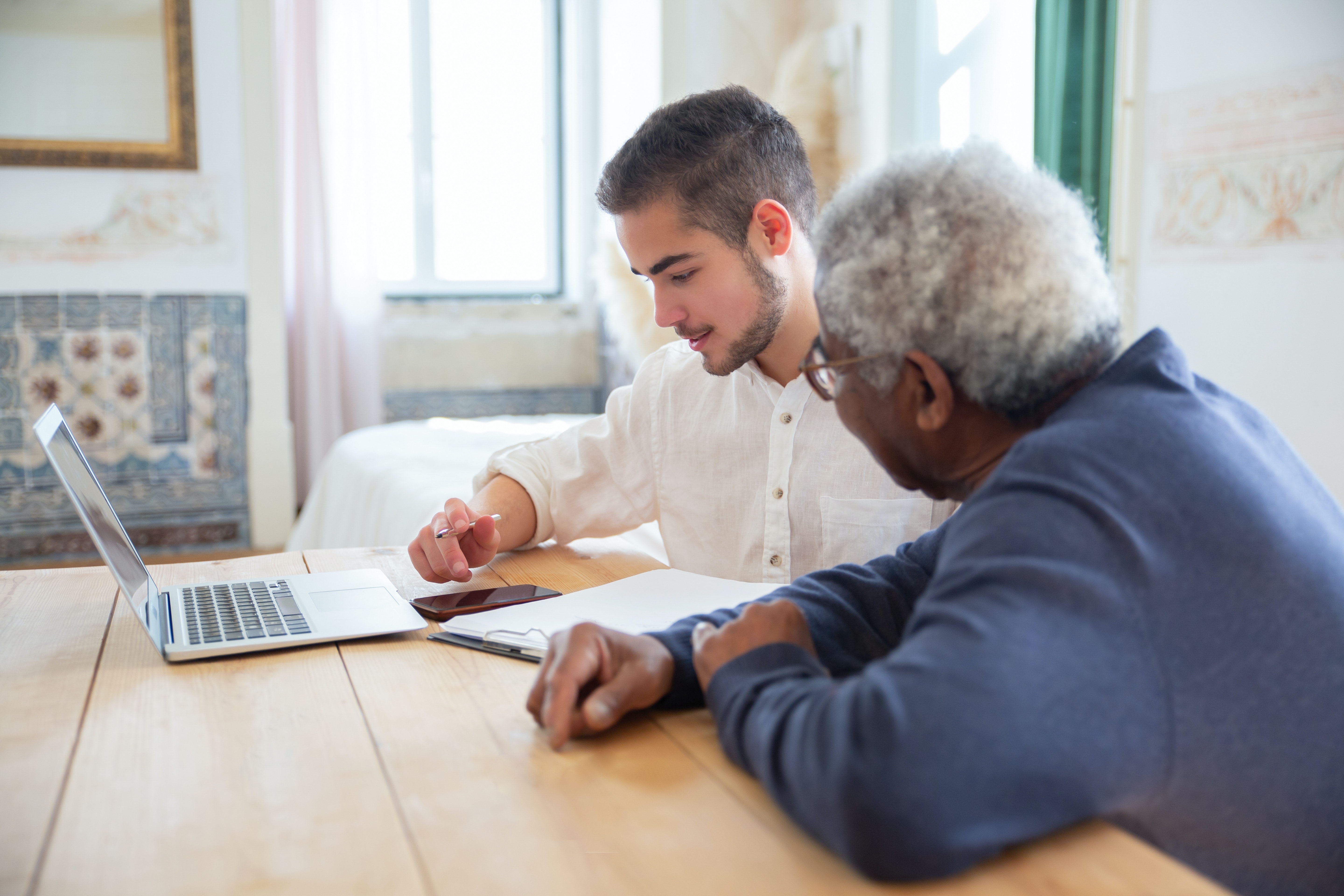 Young man discussing documents with an elderly person at a table with a laptop, a situation that could pertain to the legal distinctions between nursing home abuse and neglect.