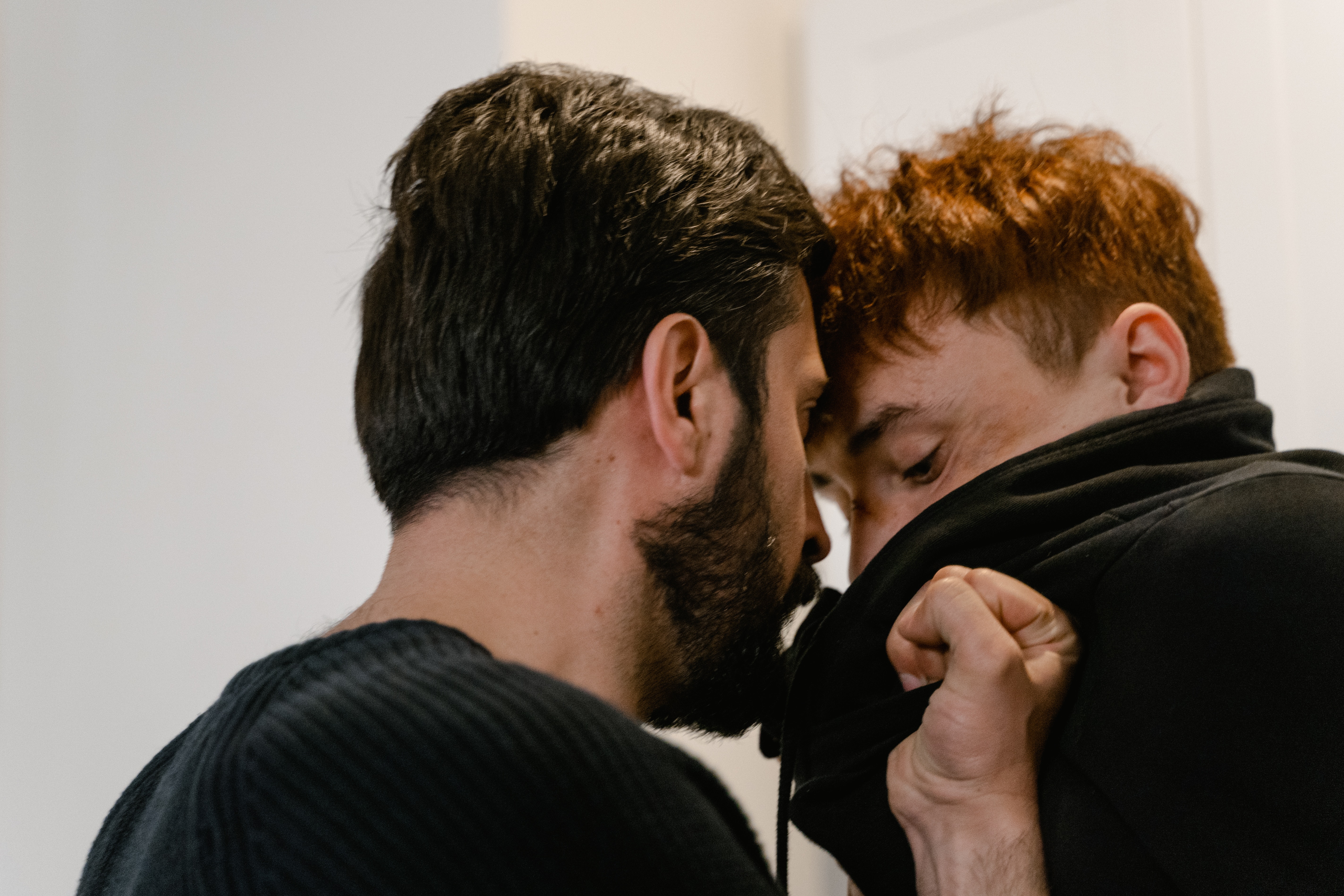 Two individuals in a close confrontation, with one appearing to whisper or speak intensely into the ear of the other who seems to be recoiling slightly. Both are partially obscured by the collar of a black jacket, indicating a potential struggle or aggressive interaction in an indoor setting with a plain white wall in the background.