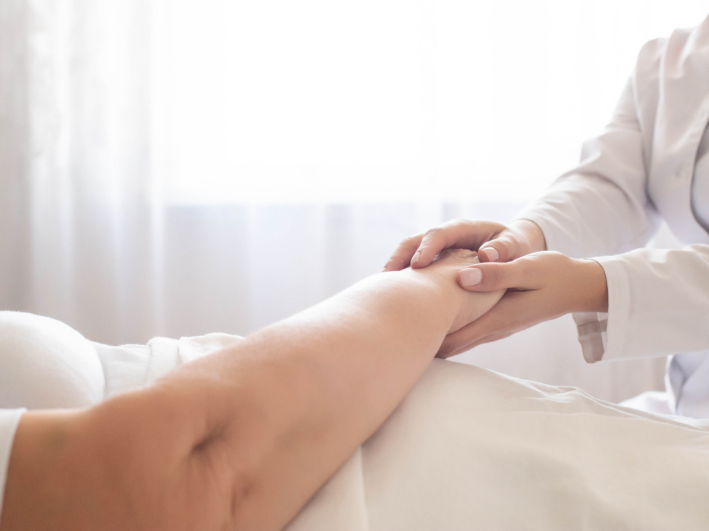 A healthcare provider in a white coat gently examines a patient's leg on a bed with soft lighting in the background, possibly evaluating for skin integrity or signs of neglect such as bedsores.