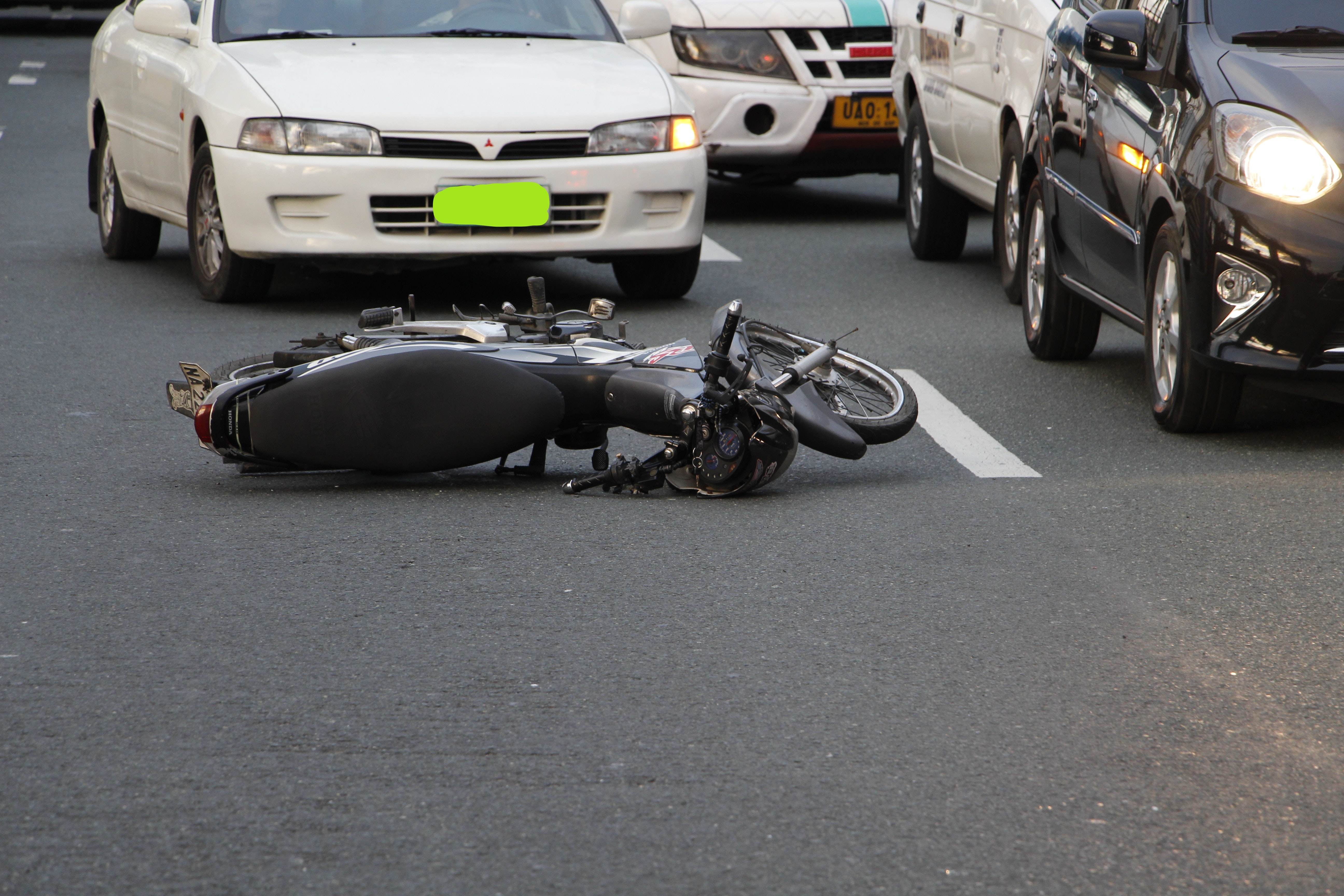 A motorcycle lies on its side in the middle of an asphalt road, with various cars halted around it, indicating a recent bike accident. The scene captures a common urban traffic incident, with the focus on the motorcycle's position, suggesting a disruption in the flow of traffic.