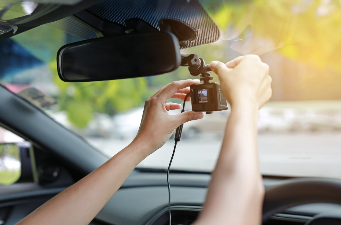 A pair of hands adjusting a dash cam attached to a vehicle's windshield, viewed from the car's interior. The camera's screen is visible, displaying the view ahead, capturing real-time driving conditions which could be critical evidence in traffic disputes or incidents.