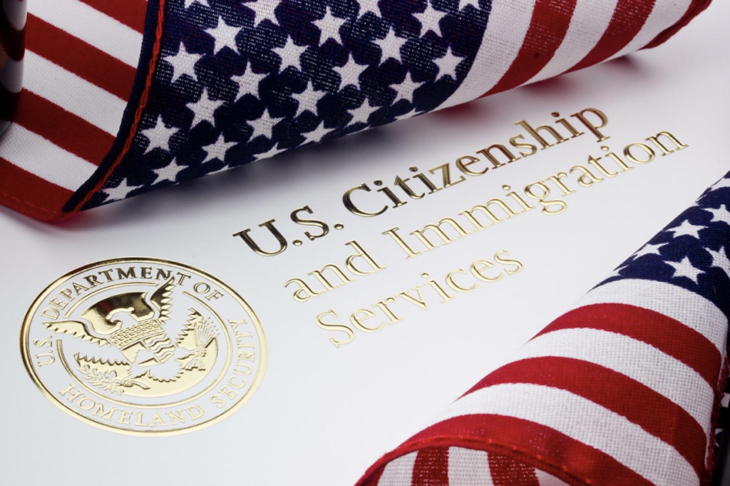 A close-up image featuring the golden seal of the U.S. Department of Homeland Security, along with the text 'U.S. Citizenship and Immigration Services' embossed in gold, alongside part of the American flag, symbolizing the immigration process in the United States.