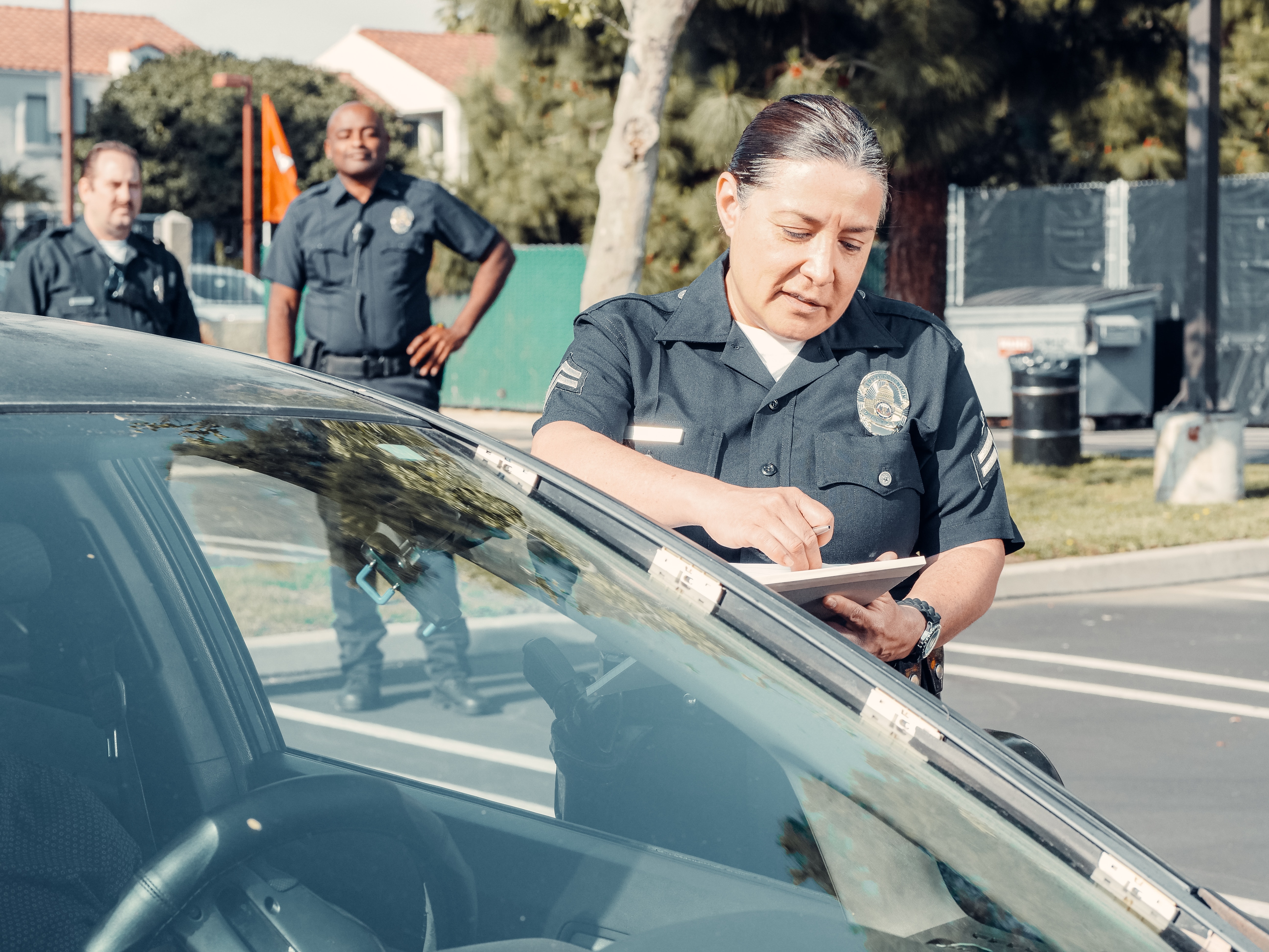A female police officer, with a badge visible on her uniform, is writing on a notepad beside a car, with two other officers standing in the background. This scene typically represents a traffic stop by law enforcement, where documentation and vehicle registration details are being checked.