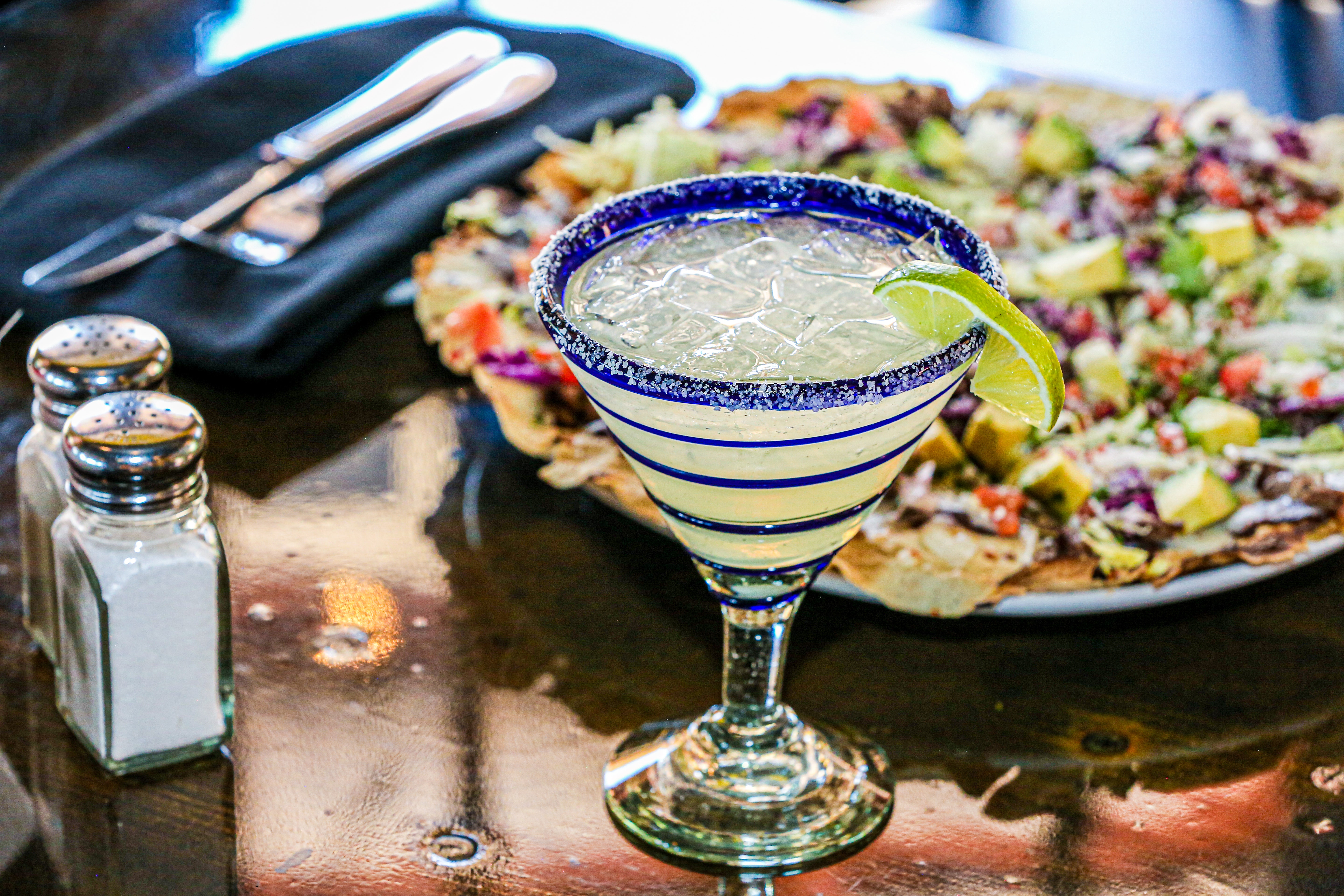 A festive margarita glass with a salted rim and lime wedge in the foreground, with a plate of nachos and condiments in the background, set on a reflective table surface, evoking a celebratory atmosphere likely related to Cinco de Mayo.