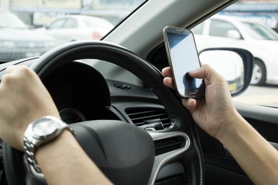 Driver's hands, one on the steering wheel and the other holding a smartphone, implying the risk of distracted driving, with traffic visible through the windshield.