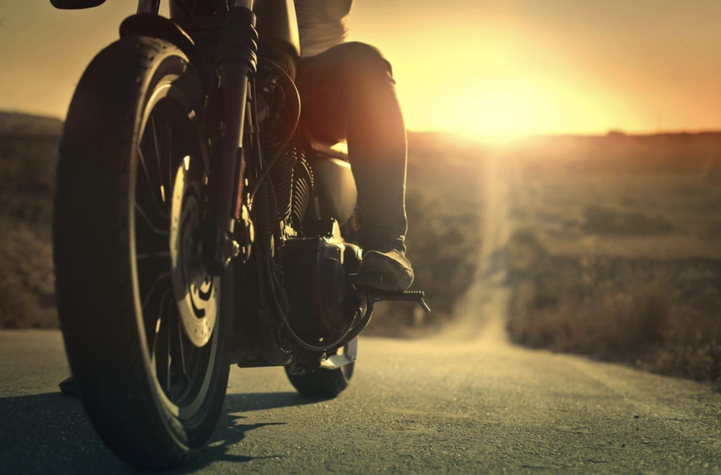 Close-up of a motorcyclist's boots on the foot pegs of a bike, with the focus on the front wheel, set against a backdrop of a sunlit open road, suggesting the importance of defensive driving for motorcyclists.