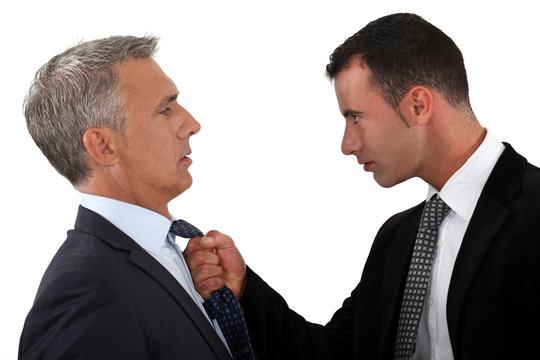 Two businessmen in suits, one appearing to forcefully grasp the tie of the other, suggesting a confrontation that may illustrate the legal concepts of duress and necessity in a professional setting.