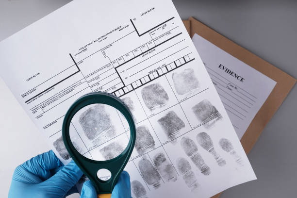 A pair of gloved hands holding a magnifying glass over a fingerprint record sheet, with an 'EVIDENCE' envelope in the background, related to the process of obtaining or reviewing fingerprint clearance cards.