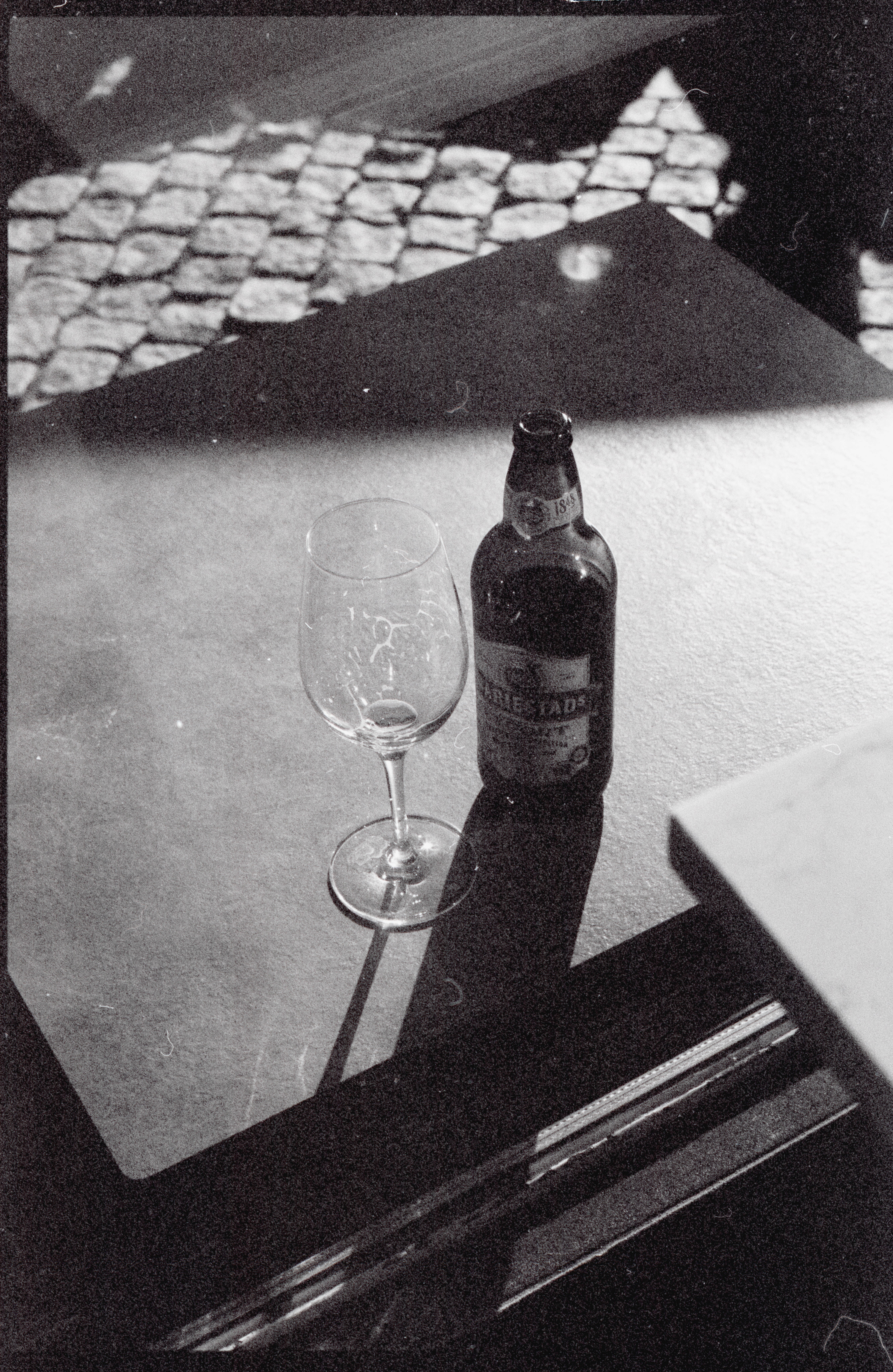 Black and white photograph of an empty wine glass and a closed bottle of beer on a reflective surface, with shadows and light creating patterns in the background, relevant to a blog post about the repercussions of DUI charges on a driver's license in Arizona.