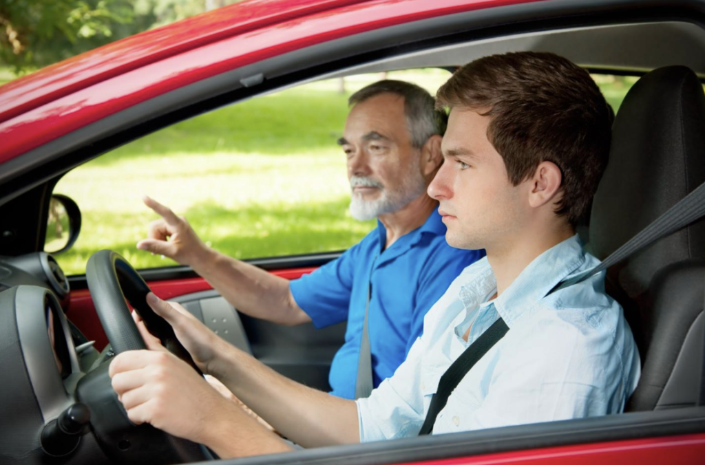 A focused teenager at the wheel of a red car with an older adult giving driving instructions, symbolizing driver's education for safe driving practices.